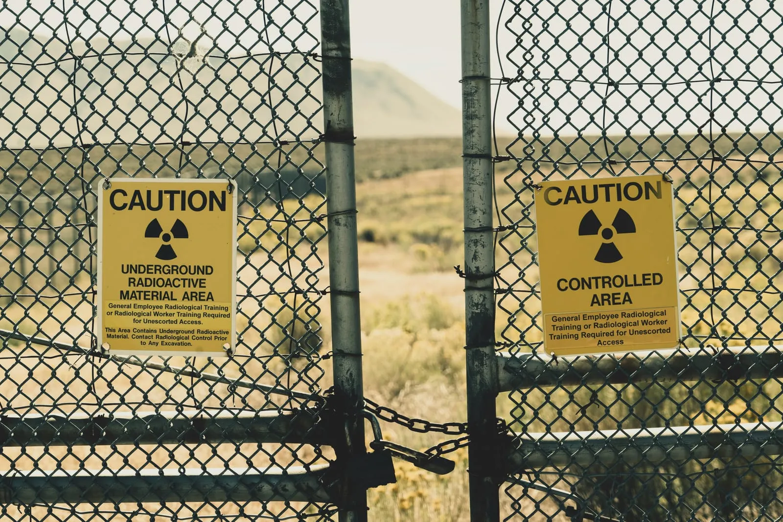 Two yellow radiation warning signs wired to a padlocked chain-link gate, reading "Caution: Underground Radioactive Material Area" and "Caution: Controlled Area". Dry scrubland and a bare hill lie behind the fence.